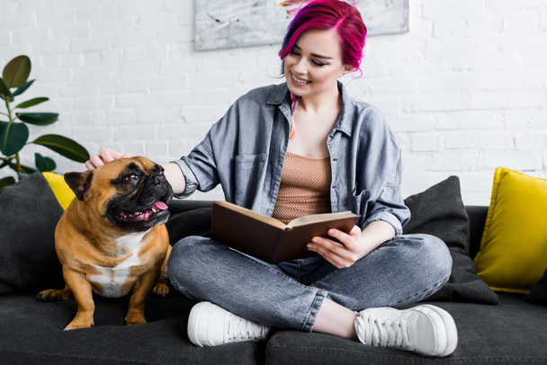 schönes Mädchen mit bunten Haaren sitzt auf dem Sofa mit Buch und streichelt süße Bulldogge - Foto, Bild