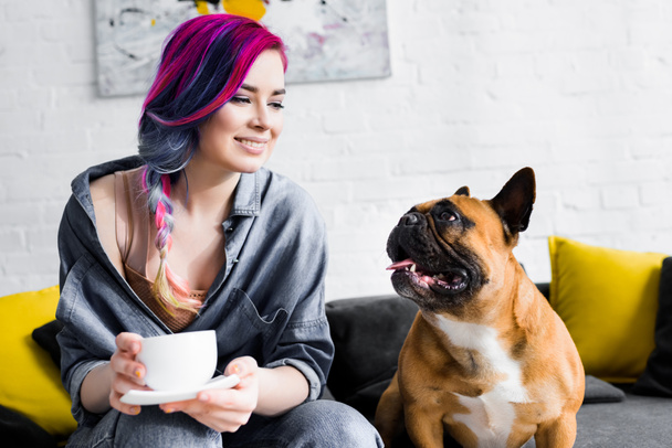 attraktives Mädchen mit buntem Haar, das eine Tasse Kaffee hält und auf einem Sofa in der Nähe einer französischen Bulldogge sitzt - Foto, Bild