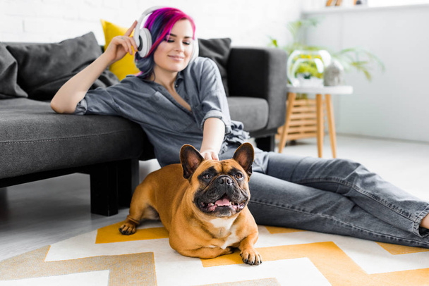 beautiful girl with colorful hair and headphones sitting on floor and petting french bulldog - Фото, изображение