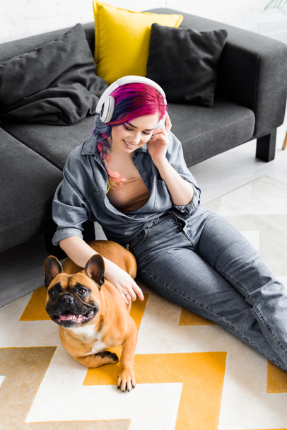 high angle view of beautiful girl with colorful hair and headphones sitting on floor and petting french bulldog - Zdjęcie, obraz
