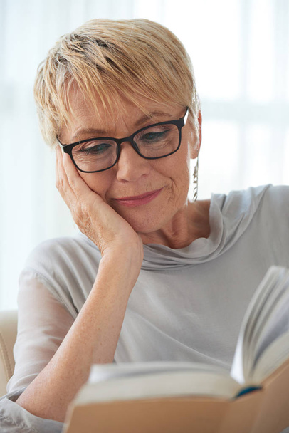 Volwassen mooie vrouw dragen bril enthousiast over het lezen van een interessant boek thuis - Foto, afbeelding
