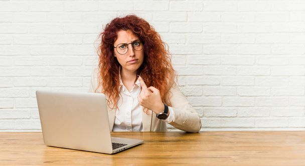 Young redhead curly woman working with her laptop pointing with finger at you as if inviting come closer. - 写真・画像