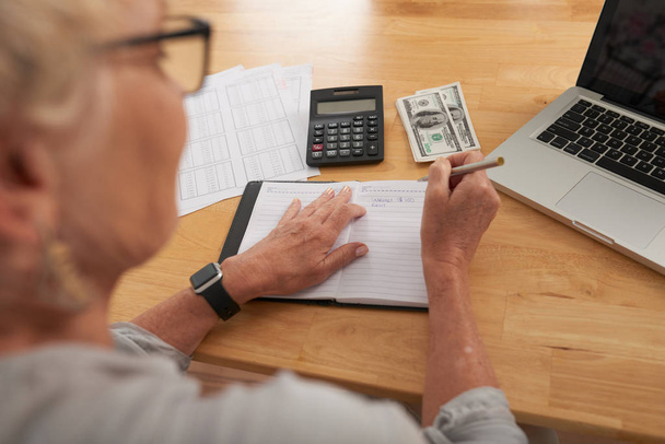 Mujer mayor sentada en la mesa con dinero y calculadora planeando su presupuesto y escribiéndolo en bloc de notas
 - Foto, Imagen