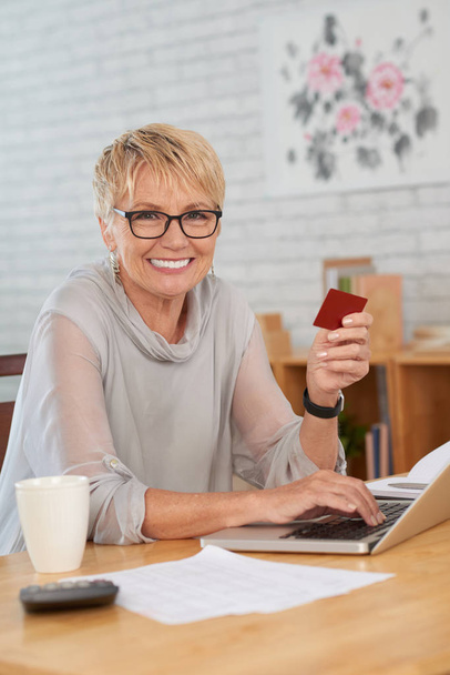 Portrait d'une femme mûre assise à la table devant un ordinateur portable et faisant des achats en ligne avec sa carte de crédit
 - Photo, image