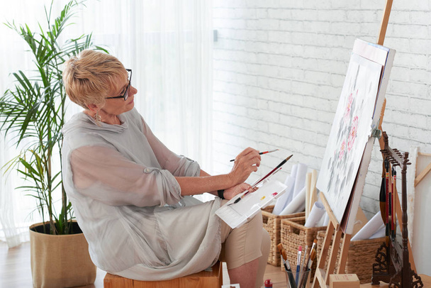 Mature woman artist sitting and painting a picture on canvas in art studio - Photo, Image