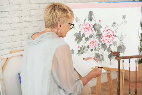 Rear view of mature woman sitting and painting a beautiful colorful picture of flowers on canvas in art studio - 写真・画像