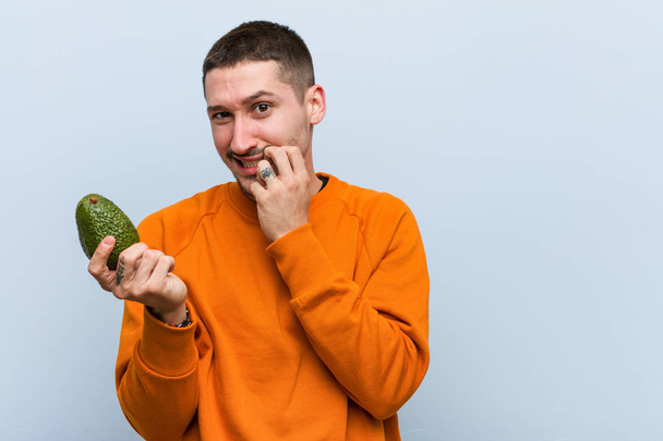 Jovem caucasiano segurando um abacate mordendo unhas, nervoso e muito ansioso
. - Foto, Imagem