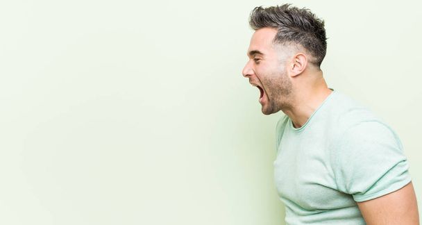 Jonge knappe man tegen een groene achtergrond schreeuwen naar een kopieerruimte - Foto, afbeelding