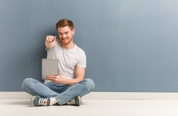 Young redhead student man sitting on the floor cheerful and smiling pointing to front. He is holding a tablet. - Photo, Image