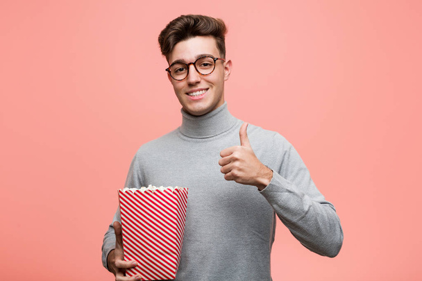 Молодий інтелектуальний чоловік тримає відро з попкорном, показуючи знак перемоги і посміхаючись широко
. - Фото, зображення