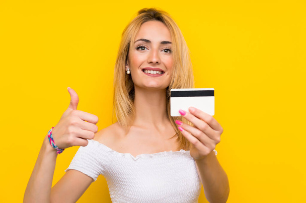 Молодая блондинка над изолированной желтой стеной держит кредитку
 - Фото, изображение