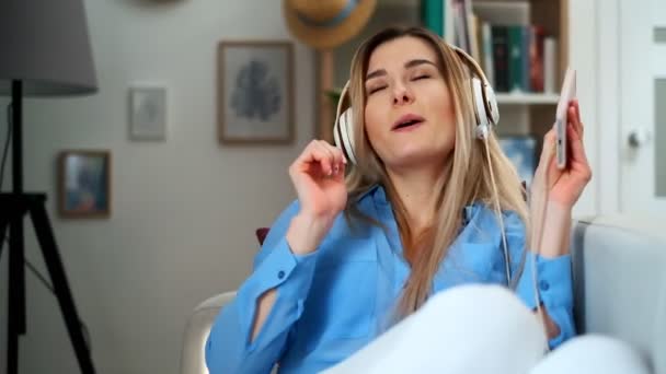 Young happy beautiful girl in blue outfit enjoying the music at home. Woman listening music in headphones from smartphone and dancing in living room. Indoor, lifestyle - Video