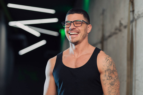 Mladý silný muž v tělocvičně v brýlích a tanktop smích - Fotografie, Obrázek