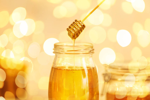 Honey dripping from dipper into jar against blurred lights, closeup - Zdjęcie, obraz