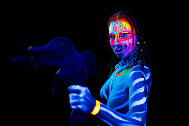 Portrait of Young naked bodyarted savage woman in blue glowing ultraviolet paint with primeval prehistorical tomahawk weapon. Agressive avatar warrior amazon with pigtails hairstyle - Photo, Image