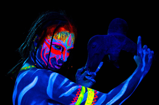 Porträt einer jungen nackten, körperbetonten Wilden in blau leuchtender ultravioletter Farbe mit urzeitlicher prähistorischer Tomahawk-Waffe. Aggressive Avatar Krieger Amazone mit Zöpfen Frisur - Foto, Bild