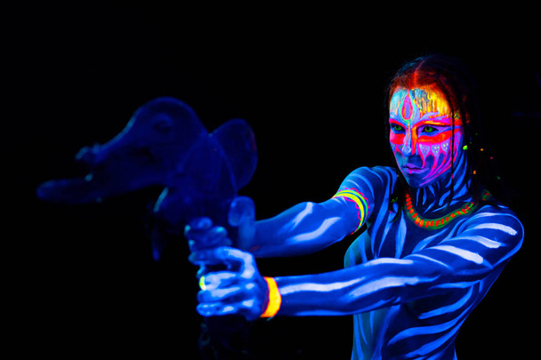 Portret młodego nagie bodyarted dziki Kobieta w niebieskim świecące ultrafioletowe farby z pierwotnej prehistorycznej broni Tomahawk. Agressive avatar wojownik Amazonka z warkoczami fryzura - Zdjęcie, obraz