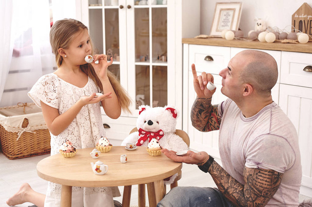 Adorable daughter wearing a white dress whith her loving father. They are drinking tea from a toy dishes in a modern kids room. Happy family. - Photo, Image