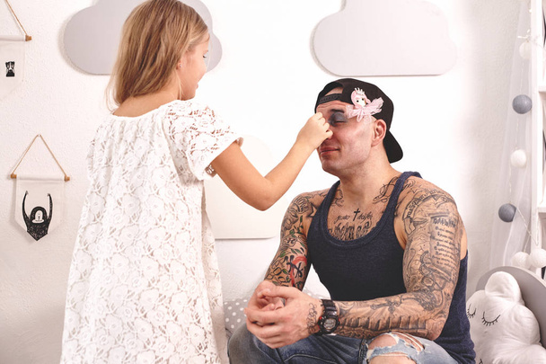 Забавно время Таттод отец в кепке и его ребенок играют дома. Милая девушка делает макияж своему отцу в своей спальне. Семейный праздник вместе
 - Фото, изображение