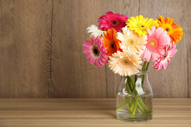 Bouquet of beautiful bright gerbera flowers in glass vase on table against wooden background. Space for text - Fotoğraf, Görsel