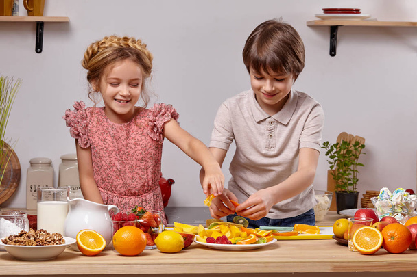 Niedliche Kinder kochen zusammen in einer Küche vor einer weißen Wand mit Regalen darauf. - Foto, Bild