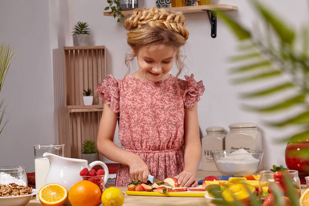 Schattig mooi meisje met een modern kapsel is koken in een keuken tegen een witte muur met planken op het. - Foto, afbeelding