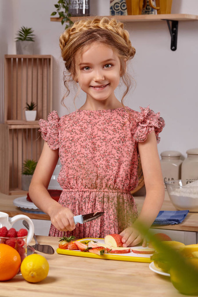 Cute beautiful girl with a modern hairstyle is cooking at a kitchen against a white wall with shelves on it. - Photo, Image