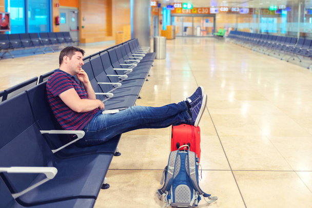 Zmęczony średnim wieku człowiek śpi z nogami na walizkę w hali odlotów, turysta śpi ze względu na opóźnienie lotu i problemy z transportem, pasażer w terminalu lotniska przy drzemkę czeka jego samolot - Zdjęcie, obraz