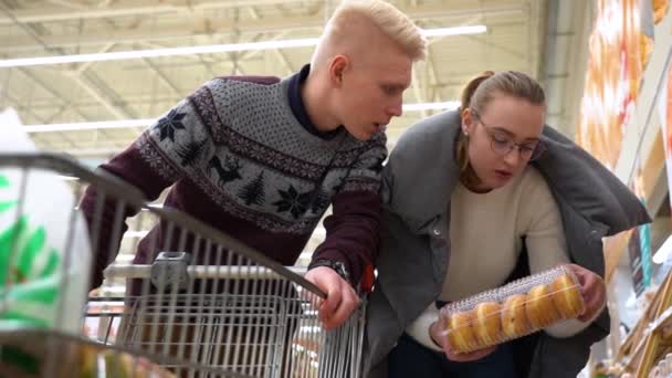 Familie paar kiest product in de hypermarkt. Jong mooi meisje met blond haar in ronde glazen in een witte wollen trui en man in grijze pullover met winkelwagen winkelen koopt donuts in de winkel. - Video