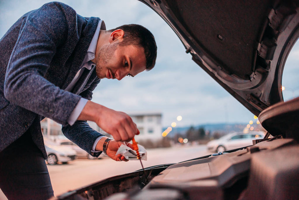 Jonge manager formeel gekleed zijn auto-olie controleren bij zonsondergang terwijl leunend onder de motorkap. - Foto, afbeelding