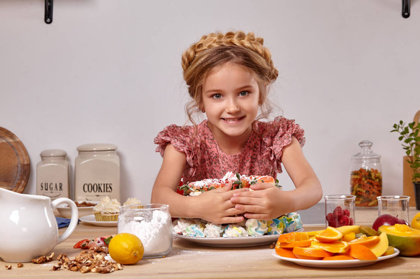 kleines Mädchen macht einen hausgemachten Kuchen mit einem einfachen Rezept in der Küche vor einer weißen Wand mit Regalen darauf. - Foto, Bild