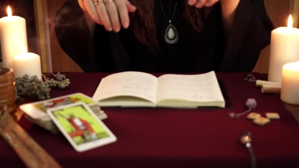 Čarodějka je věštkyně v černém plášti, drží si ruce nad knihou, čte kouzlo a volá duchy. Tarot karty, bílé svíčky a starověké runy na mystickém pozadí. Koncepce okultismu, esoterického a jasnovidectví - Záběry, video