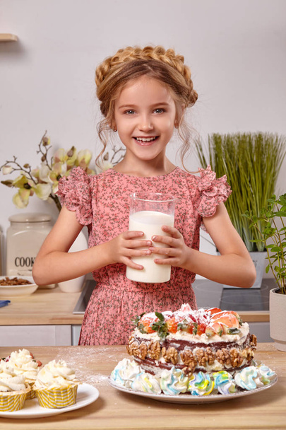 Pikkutyttö tekee kotitekoista kakkua, jossa on helppo resepti keittiössä valkoista seinää vasten, jossa on hyllyt.
. - Valokuva, kuva