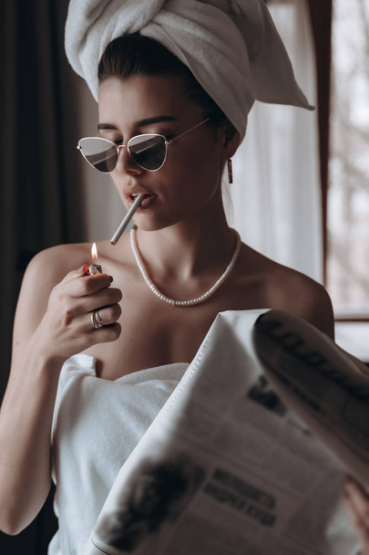 Όμορφη νεαρή γυναίκα με πετσέτα καπνίζει τσιγάρο και διαβάζει εφημερίδα. - Φωτογραφία, εικόνα