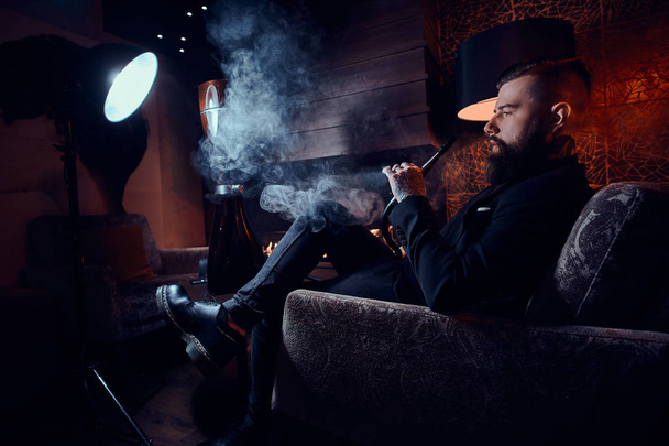 Ο μουσάτος χαλαρός άνθρωπος κάθεται στην πολυθρόνα και καπνίζει χουλέ κοντά στο τζάκι - Φωτογραφία, εικόνα