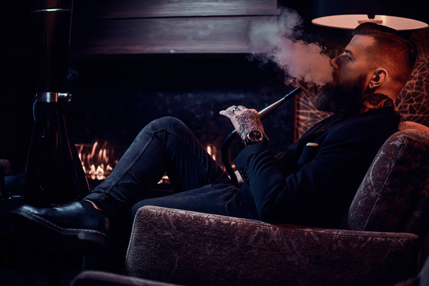 Bearded relaxed man is sitting on the armchair and smoking hookah near fireplace - Фото, изображение