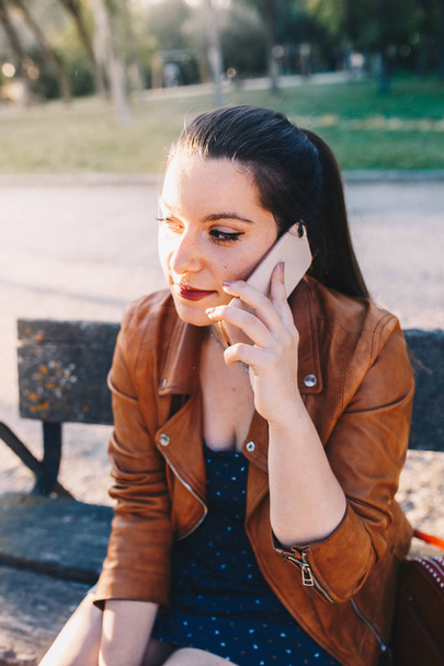 Mujer joven feliz con chaqueta de cuero hablando y escuchando en el teléfono inteligente teléfono inteligente en un parque de la ciudad sentado en un banco
 - Foto, imagen