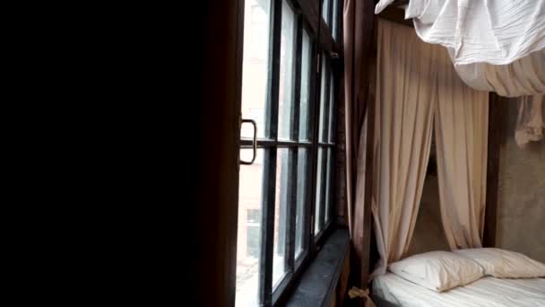 Een blik uit de donkere hoek van de slaapkamer in retro-stijl met wit bed met gordijnen en twee kussens op de achtergrond van beige stenen muur naast de oude heldere zwarte houten ramen. - Video