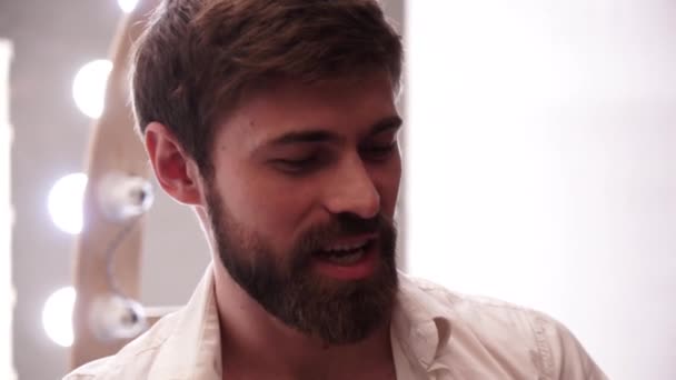 Portrait of a brutal Caucasian bearded man in a white unbuttoned shirt. A man growls and smiles looking at the camera against the background of a white wall and bright LED lights and light bulbs. - Materiaali, video