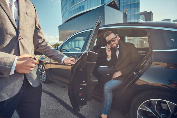 Atrakcyjny mężczyzna w okularach jest rozmowa przez smartphone i siedzi w samochodzie, podczas gdy jego asystent jest otwarcie drzwi dla niego. - Zdjęcie, obraz