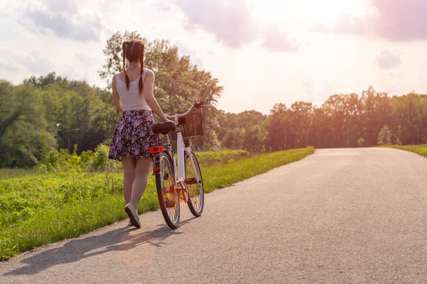 市内の公園の道路上の夏の夕日に自転車を持つ女の子。ぼやけた夏の背景にサイクルクローズアップホイール。夏の日没時に働くために通りをサイクリング。自転車とエコロジーライフスタイルのコンセプト. - 写真・画像