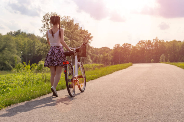 Girl with bike at the summer sunset on the road in the city park. Cycle closeup wheel on blurred summer background. Cycling down the street to work at summer sunset. Bicycle and ecology lifestyle concept. - Valokuva, kuva