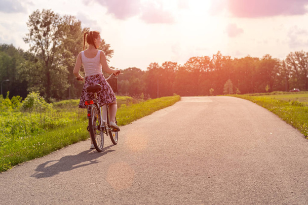 Şehir parkında yolda yaz gün batımında bisikletli kız. Bulanık yaz arka planında yakın çekim tekerleği devir. Yaz gün batımında çalışmak için sokakta bisiklete binmek. Bisiklet ve ekoloji yaşam tarzı kavramı. - Fotoğraf, Görsel