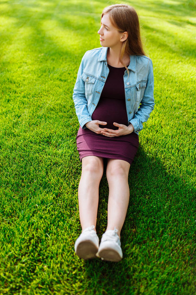 Счастливая молодая беременная женщина сидит в парке на газоне
, - Фото, изображение
