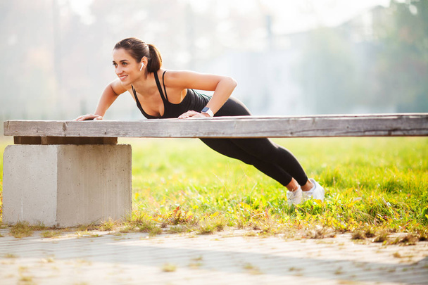 Portrait of beautiful sporty woman 20s in sportswear doing push-ups, and listening to music with bluetooth earbud during workout in green park - Foto, Imagen