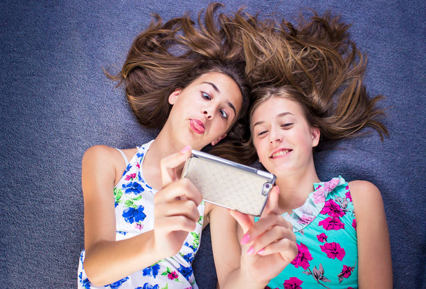 Two girls lying on the bed looking at smart phone, taking selfie being goofy. - Fotoğraf, Görsel