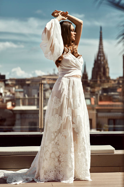 Young bride in white wedding dress posing with rising hands outdoor on city background - Photo, Image