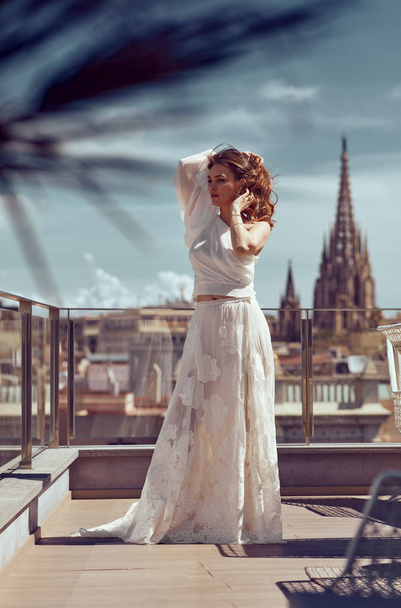 Young bride in white wedding dress posing outdoor, playing with hair over city view - Фото, изображение
