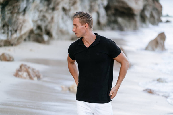 Sexy muscular man in black t-shirt resting on beach, ocean waves at background.  - Φωτογραφία, εικόνα
