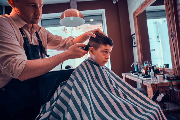 Cute little child is getting trendy haircut from barber at busy barbershop - Photo, Image